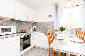 A modern kitchen with white cabinets, granite countertops, a built-in oven, a microwave, and a dining area with a table set for four. - Cozy vacation apartment on the 1st floor in a quiet location in Wenningstedt (Wenningstedt)