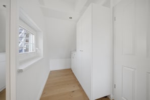 A narrow hallway with white walls and wooden flooring, leading to a door with a window on the left.