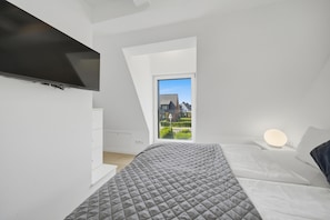 A modern bedroom with a sloped ceiling, a bed with a quilted grey duvet, a bedside table with a lamp, and a view of a house through a window.