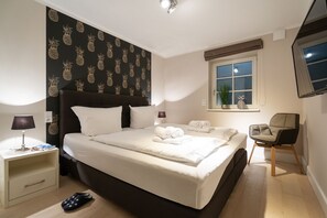 Une chambre bien rangée, avec un grand lit, des tables de chevet, une chaise et une fenêtre agrémentée d’une plante.