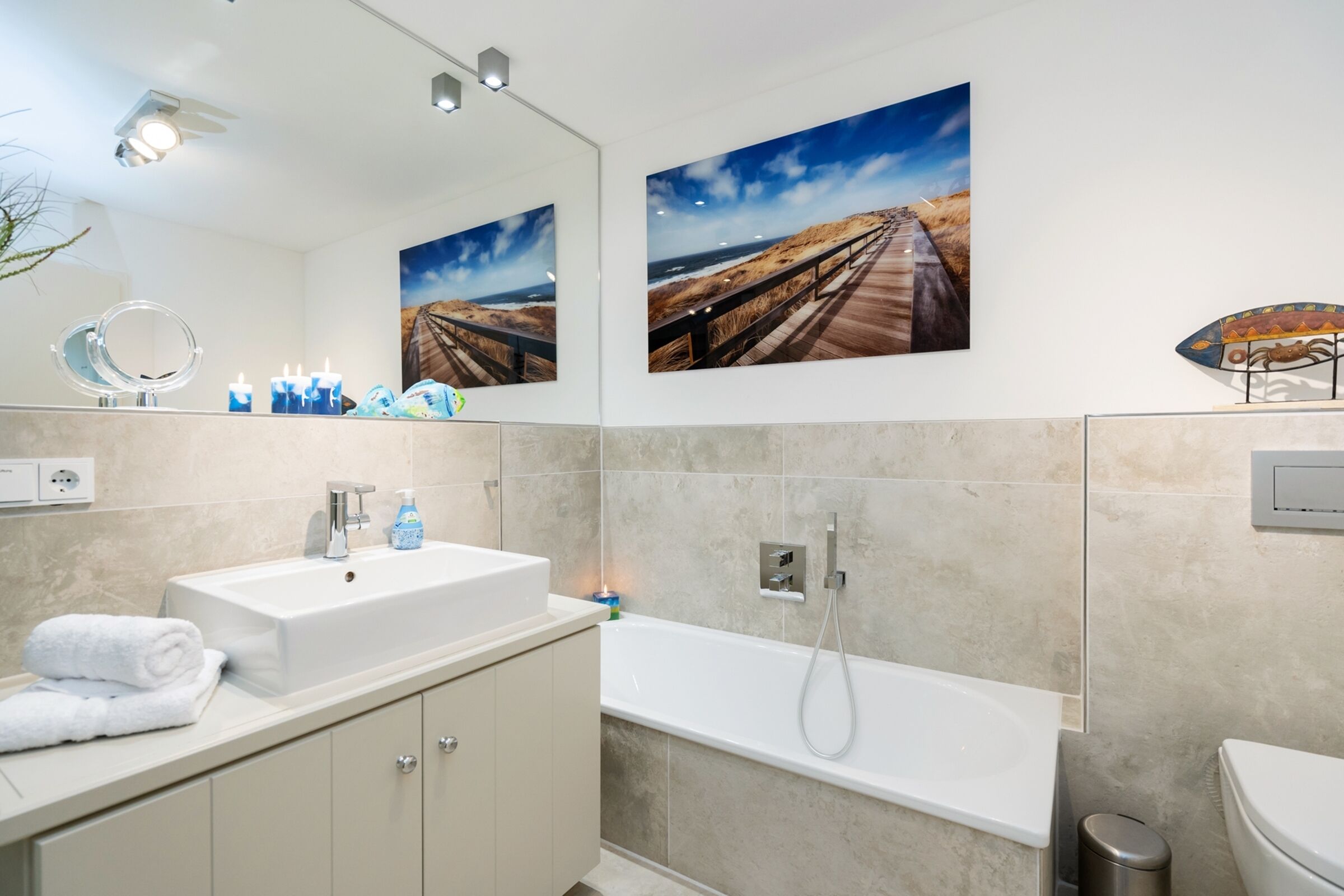 A modern bathroom with a large mirror, a sink, a bathtub, and two wall-mounted artwork depicting coastal scenes.