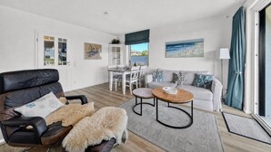 Ett modernt vardagsrum med en lädersoffa, ett vitt matbord och utsikt över havet genom ett stort fönster.