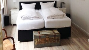 Ein Doppelbett mit weißen Leinentüchern, eine hölzerne Kommode, ein Nachttisch mit Lampe und Wandleuchten.