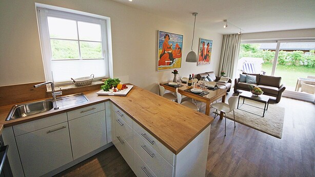 Een moderne keuken met een houten aanrecht, witte kasten en een groot raam. Er is een zithoek met een tafel, stoelen en een bank.