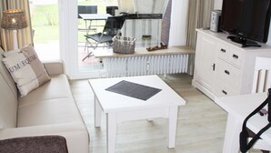 Ein modernes Wohnzimmer mit einem weißen Sofa, einem braunen Sessel, einem weißen Couchtisch, einem Fernseher auf einem Schrank und Blick ins Freie durch große Glasüren.