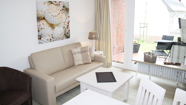 Ein modernes Wohnzimmer mit einem beigen Sofa, einem weißen Couchtisch und einem großen Fenster mit Blick auf einen Garten und eine Ziegelmauer.