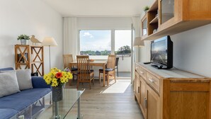 Ein modernes Wohnzimmer mit Sofa, Esstisch, Stühlen, Fernseher und Blick durch ein großes Fenster.