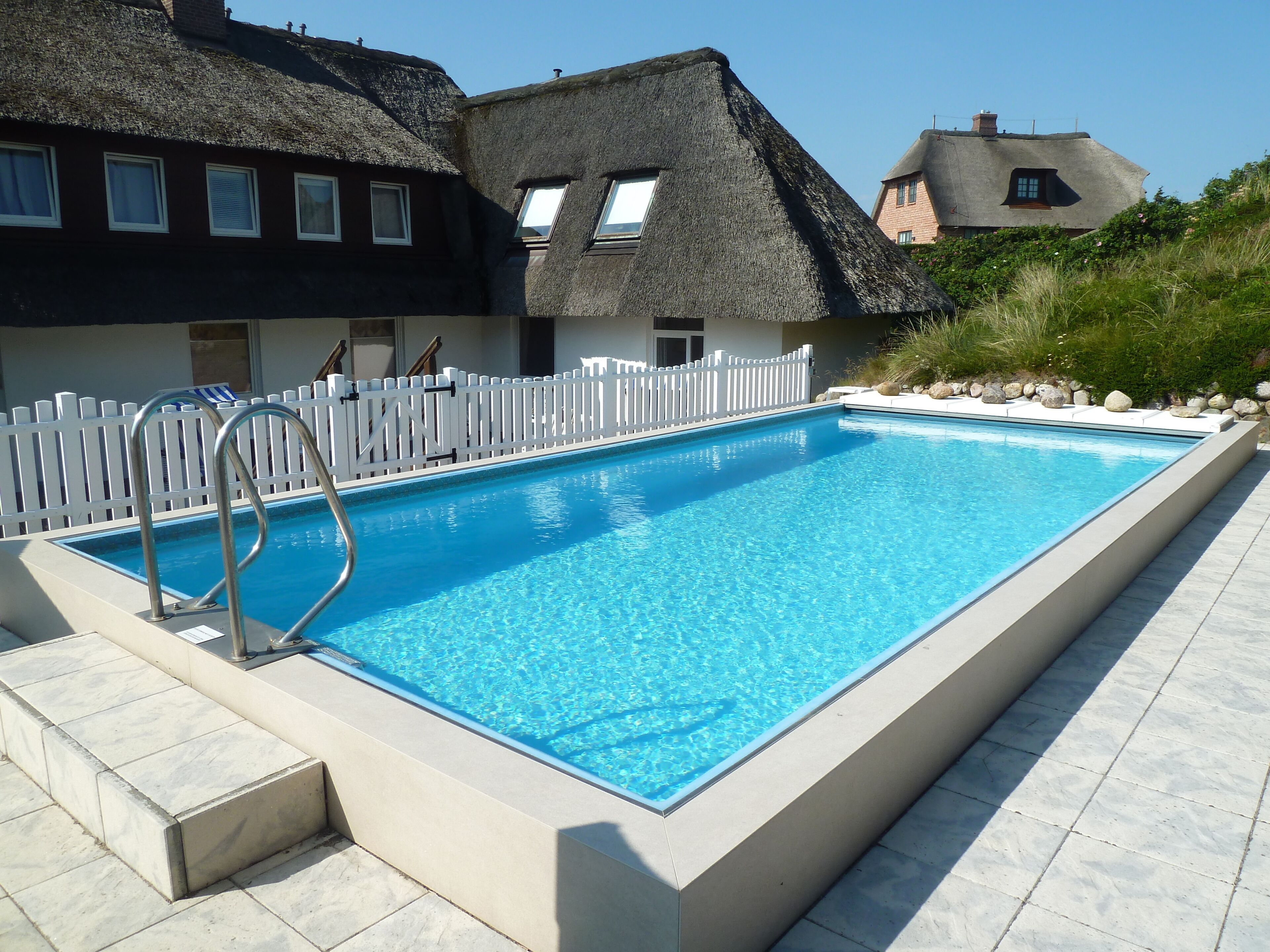 A rectangular swimming pool with a metal ladder on one side, surrounded by a white fence.