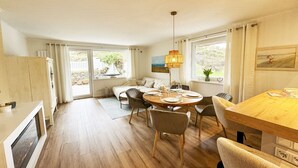 Uma área de refeições acolhedora com uma mesa de madeira posta para quatro pessoas, rodeada por cadeiras. Uma grande janela oferece luz natural, e um sofá é visível ao fundo.