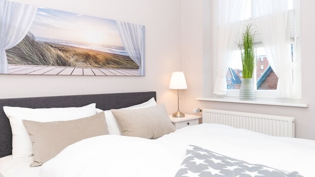 A neatly made bed with a star-patterned blanket, a nightstand with a lamp, a window with a plant, and a beach scene painting on the wall. - 3-room apartment on the upper floor in a quiet location in Alt-Westerland (Westerland)