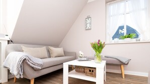A modern living room with a grey sofa, a white coffee table, and a window with a view of a blue house. - 3-room apartment on the upper floor in a quiet location in Alt-Westerland (Westerland)