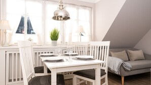 A dining area with a white table set for four, surrounded by white chairs. There is a grey sofa with a throw blanket and a potted plant on the windowsill. - 3-room apartment on the upper floor in a quiet location in Alt-Westerland (Westerland)