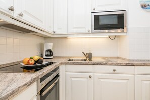 A modern kitchen with white cabinets, a built-in oven, a microwave, a granite countertop, and a fruit bowl. - Modern 2-room apartment in a quiet location in Wenningstedt (Wenningstedt)
