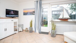 A modern living room with a television, a painting of a sunset, a sailboat model, and a view of a house through a glass door. - Comfortable house in Westerland with terrace & garden (Westerland)
