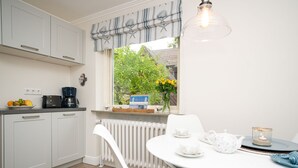 A modern kitchen with white cabinets, a dining table with white chairs, and a window with a view of a garden. - Comfortable house in Westerland with terrace & garden (Westerland)