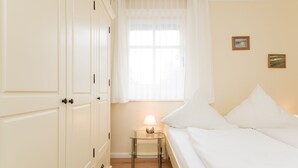 A bedroom with a bed, a nightstand with a lamp, a window with curtains, and two framed pictures on the wall. - 3-room apartment on the ground floor in a quiet location in Alt-Westerland (Westerland)