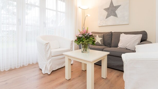 A living room with a white sofa, a grey sofa, a white armchair, and a small white table with a vase of flowers. - 3-room apartment on the ground floor in a quiet location in Alt-Westerland (Westerland)