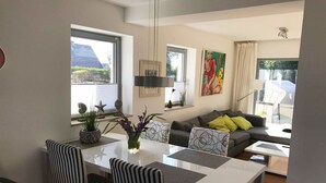 A modern living room with a dining area, featuring a sofa with patterned cushions, a dining table with chairs, and a large window providing a view of the outdoors. - Spacious 3-room apartment with terrace and garden in Wenningstedt (Wenningstedt)