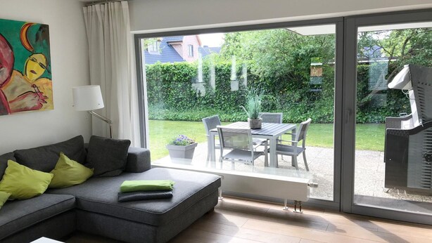 A modern living room with a grey sectional sofa, a white coffee table, and a dining area with a table and chairs. A large sliding glass door leads to a garden with a barbecue and a view of a house and greenery. - Spacious 3-room apartment with terrace and garden in Wenningstedt (Wenningstedt)