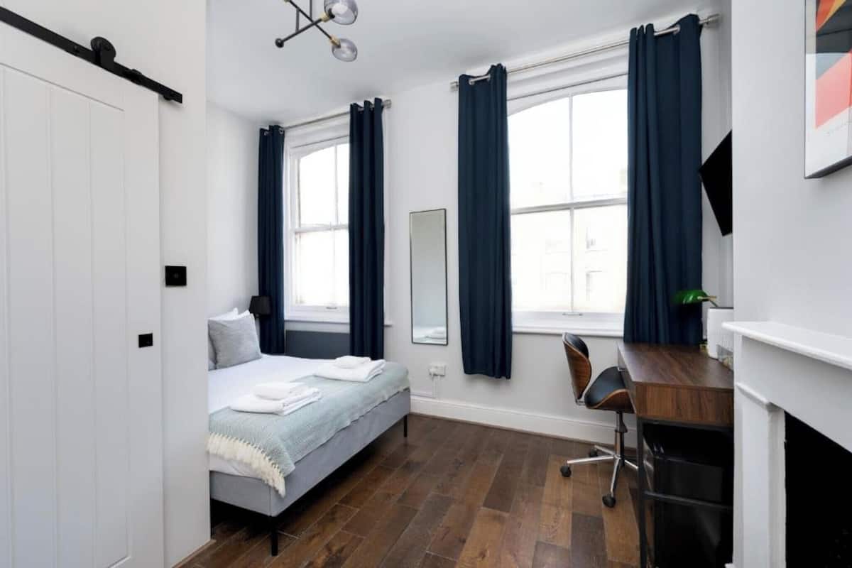 A bedroom with a bed, a desk, a chair, a mirror, and a fireplace.