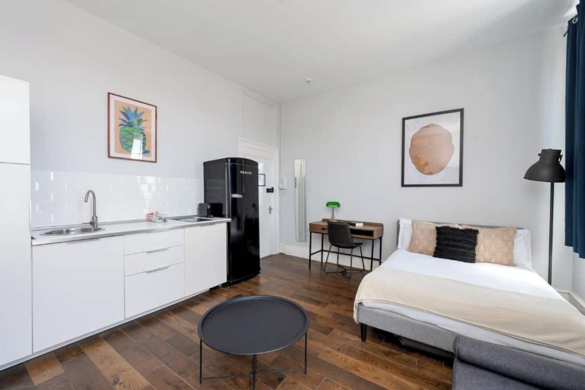 A compact hotel room with a kitchenette, a bed, a small round table, and a desk.