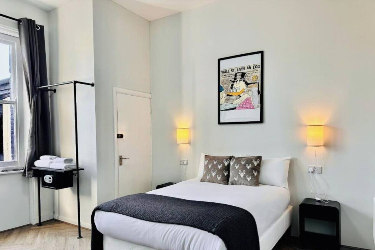 A hotel room with a bed, bedside tables, a wall-mounted light, a door, a window with curtains, and a towel rack.