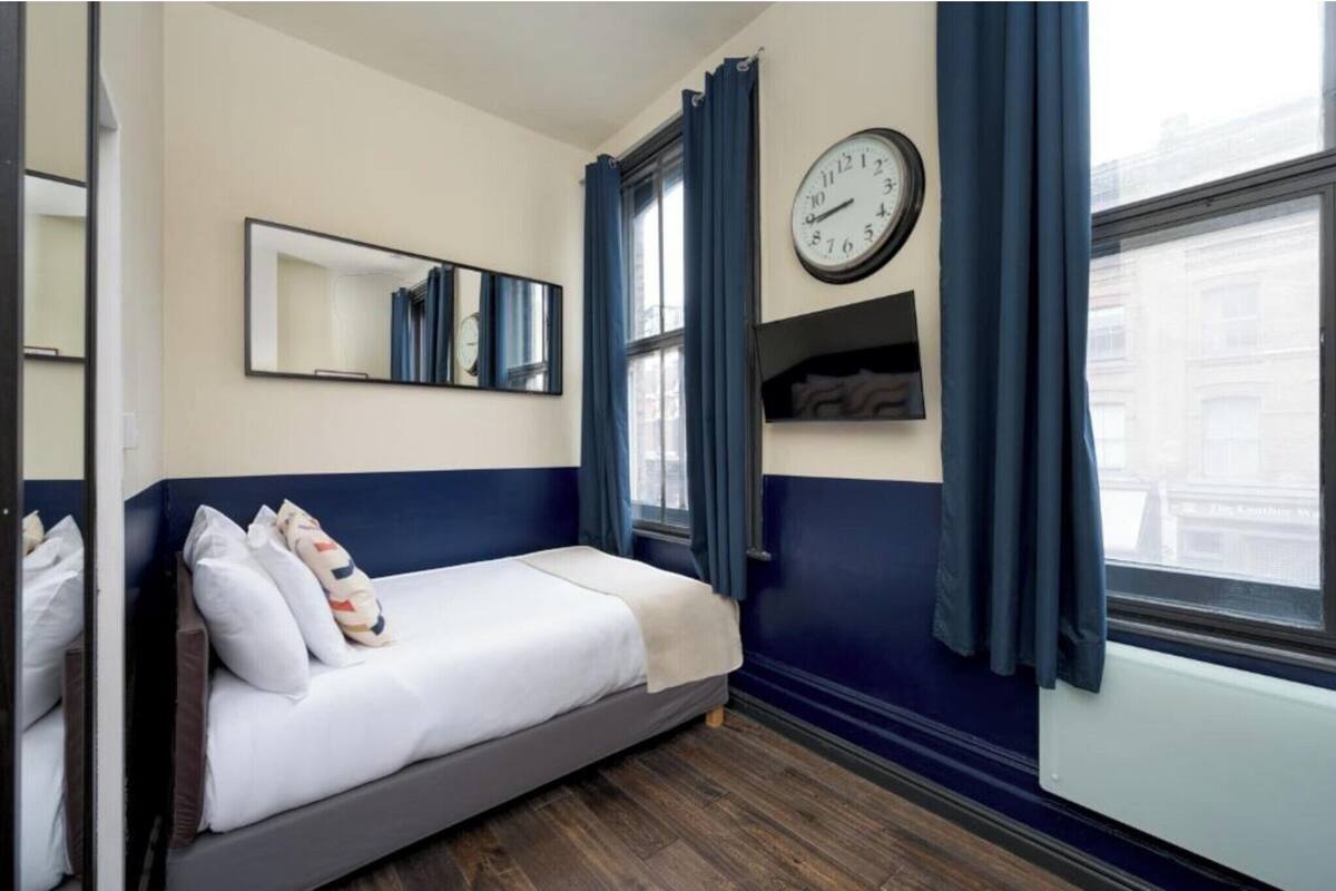 A single bed with a white mattress and pillows, a wall clock, a mirror, and a window with blue curtains.