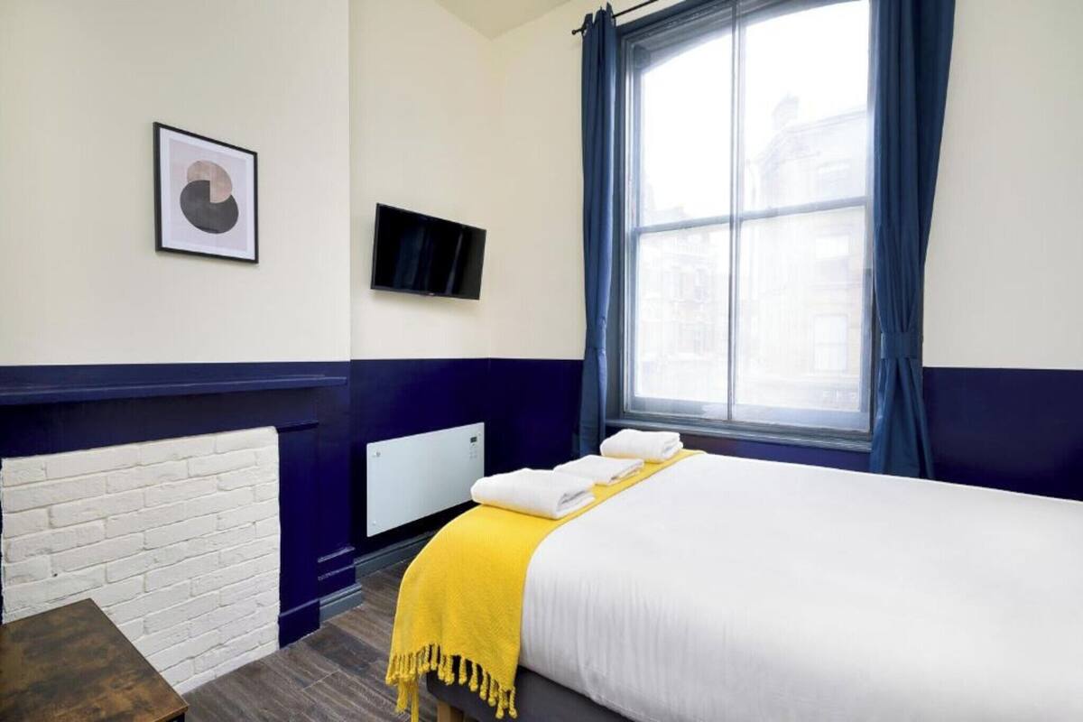 A hotel room with a bed, a white brick wall, a flat-screen TV, and a window with curtains.