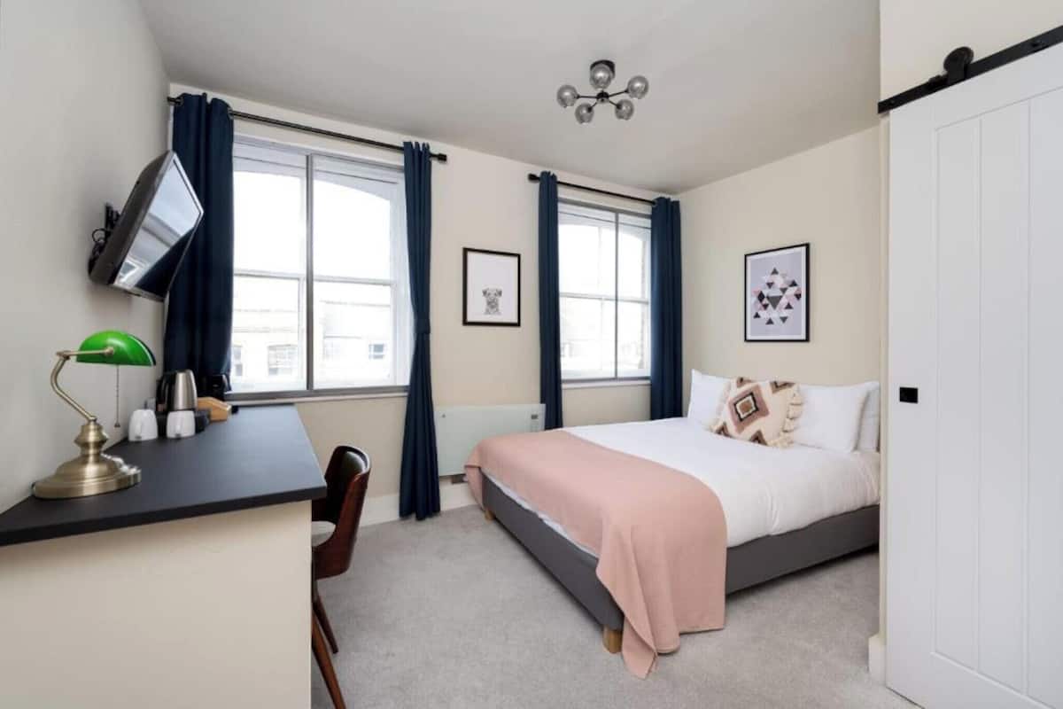 A bedroom with a bed, a desk with a lamp, a chair, and two windows with curtains.