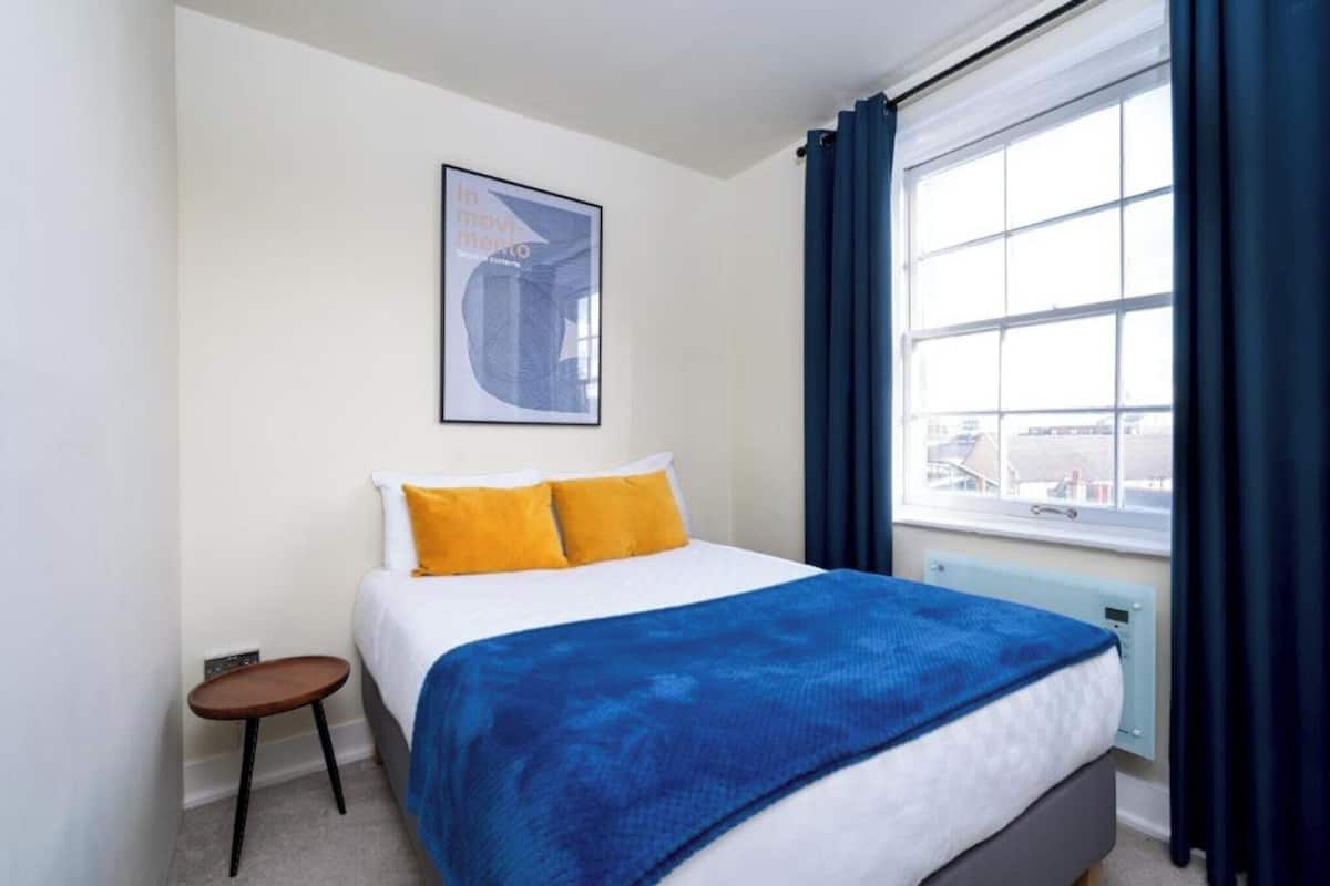 A hotel room with a bed, a small table, a window with curtains, and a framed picture on the wall.