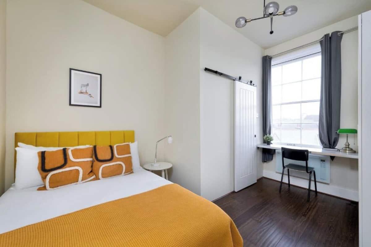 A hotel room with a bed, a desk, a chair, a window with curtains, a radiator, and a picture on the wall.