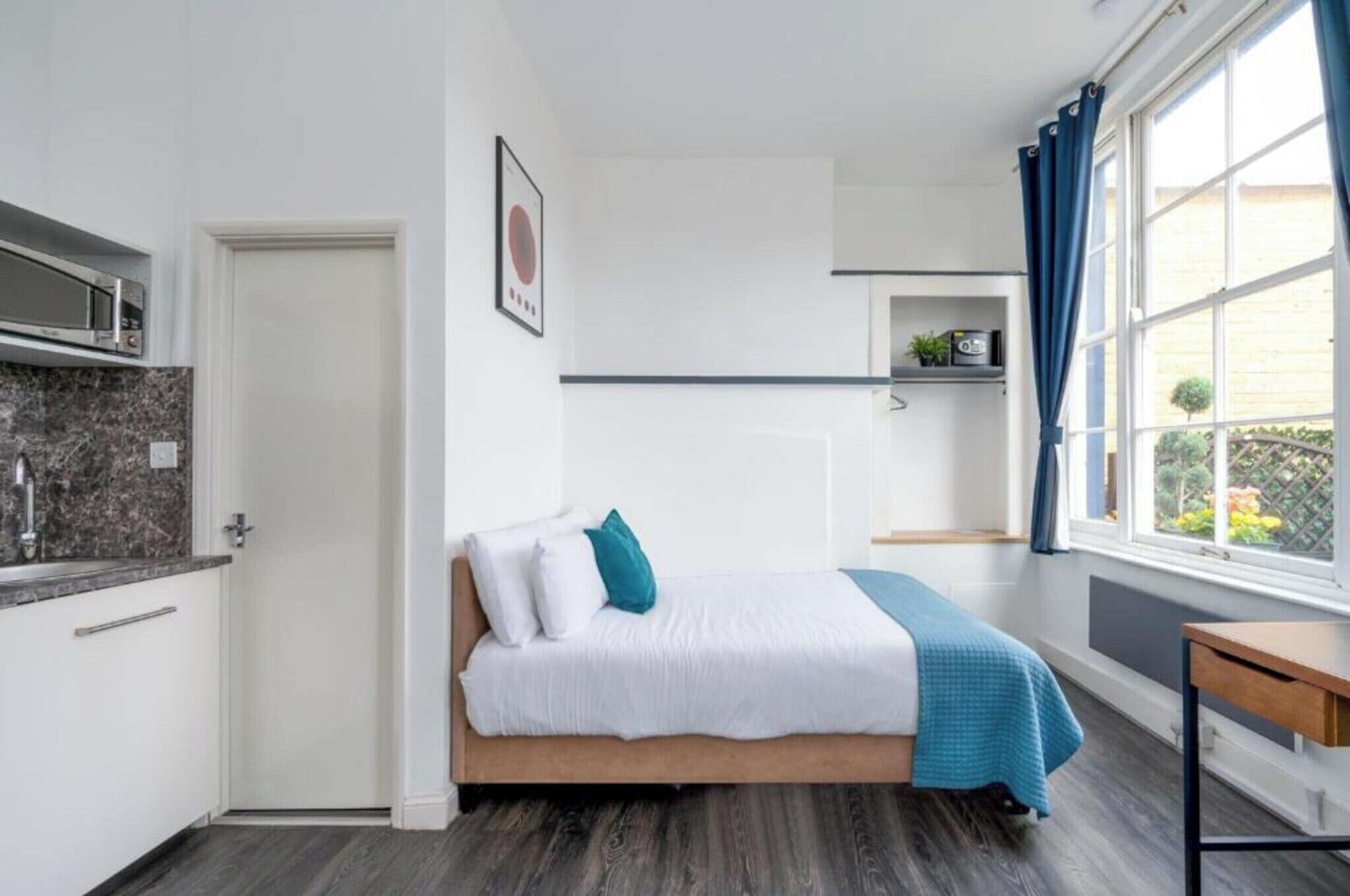 A bedroom with a bed, a window with curtains, a small table, and a kitchenette.