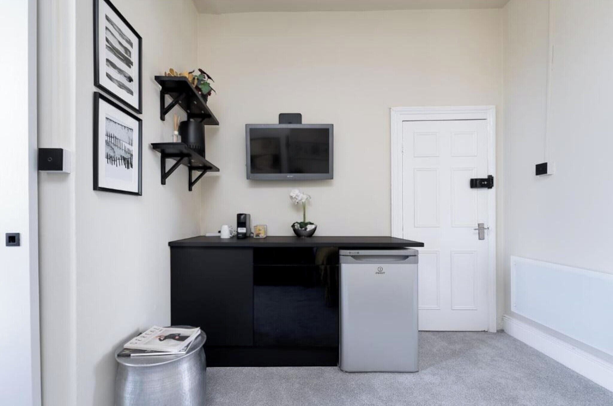 A compact room with a black desk, a small refrigerator, a white door, and framed pictures on the wall.