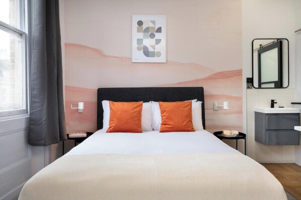 Une chambre d’hôtel moderne avec un grand lit, des oreillers orange, une table de chevet, une petite table d’appoint, un miroir fixé au mur et une petite table avec une lampe.