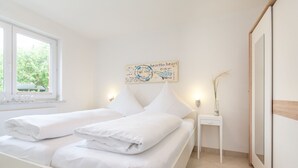 Un lit bien fait, avec du linge de lit blanc, une table de chevet ornée d’un vase et un mur sur lequel est inscrit un proverbe inspirant.