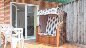 Une terrasse avec un banc en bois et une chaise en plastique blanche, une porte-fenêtre coulissante et une clôture en bois.