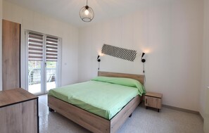 Ett sovrum med en säng i trä, ett nattduksbord, en väggmonterad högtalare och en skjutdörr i glas som leder ut till ett utomhusområde.