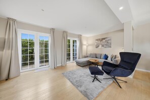 Ein geräumiges Wohnzimmer mit einem grauen Sofa, einem dunkelgrauen Sessel, einem Couchtisch und einem großen Fenster mit Vorhängen.