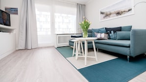 A modern living room with a grey sofa, a small white table, a green rug, and a large window with curtains. - Modern vacation apartment with sauna and heated indoor pool in Westerland (Westerland)