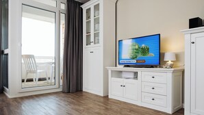 Uma sala de estar moderna com uma televisão de tela plana, um armário branco e uma porta de vidro deslizante que dá para uma varanda com uma cadeira branca.