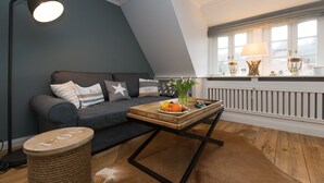 Ein modernes Wohnzimmer mit dunkelblauer Akzentwand, einem grauen Sofa mit Zierkissen, einem Holztisch mit Früchten und einer Vase, einer Stehlampe und einem Fenster mit Jalousien.