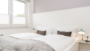 Ein Doppelbett mit weißer Bettwäsche und einer grauen, gemusterten Tagesdecke, zwei Nachttische mit Lampen, ein Fenster mit Jalousien und ein weißes Kopfteil.