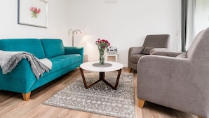 Ein Wohnzimmer mit einem blaugrünen Sofa, einem grauen Sessel, einem runden Couchtisch mit Blumen und einem gemusterten Teppich.