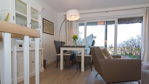A dining area with a table set for four, surrounded by chairs. There is a large window with a view of the outdoors. - Beautiful 3-room apartment with direct view of the mudflats in Rantum (Rantum)