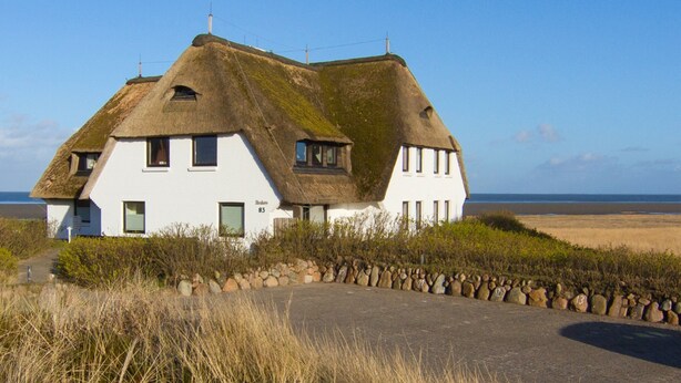 A white house with a thatched roof, situated on a grassy area with a stone wall in the foreground. - Beautiful 3-room apartment with direct view of the mudflats in Rantum (Rantum)