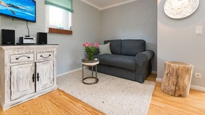 Uma sala de estar com um sofá cinza, um armário branco com caixas de som, uma televisão, uma mesinha redonda com um vaso de flores e um banquinho de madeira.