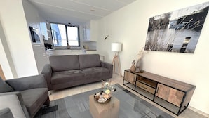 Un salon moderne avec un canapé gris, une table basse agrémentée d’objets décoratifs, une console en bois et une grande fenêtre donnant sur des immeubles.