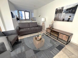 A modern living room with a grey sofa, a coffee table with decorative items, a wooden console table, and a large window with a view of buildings. - Park Brekkense Wiel 044 (Lemmer)