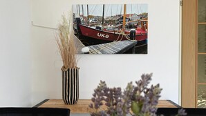 Une table à manger ornée d’un vase contenant des plantes séchées, d’une photographie encadrée représentant des bateaux et d’une porte en bois.