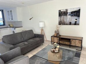 A modern living room with a grey sofa, a wooden coffee table with decorative items, a painting on the wall, and a kitchen area with a washing machine. - Park Brekkense Wiel 044 (Lemmer)